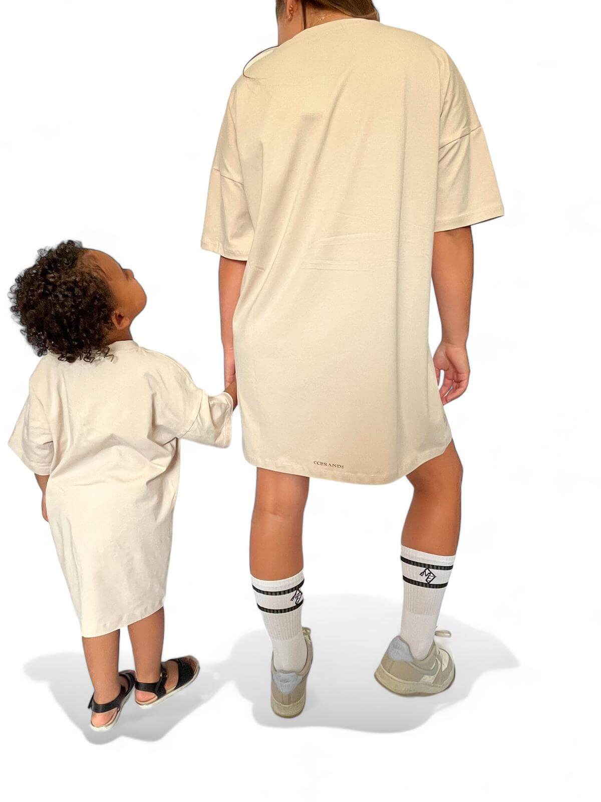 Achterkant van beige oversized T-shirt jurk met gouden opdruk 'Senorita' – vrouw hand in hand met klein meisje in dezelfde jurk.